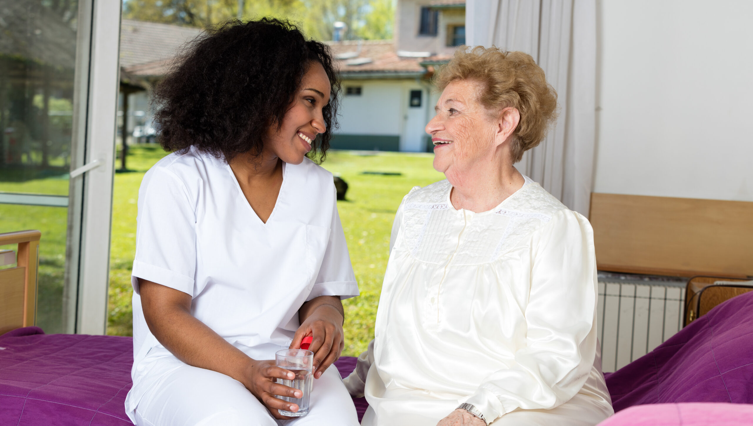 Nurse giving glass of water to elder woman seated in hospital be
